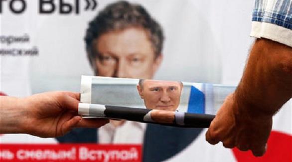 رجل يمسك بصحيفة فيها صورة الرئيس الروسي فلاديمير بوتين، أمام ملصق لقيادي ليبرالي في البلاد(إ ب أ)